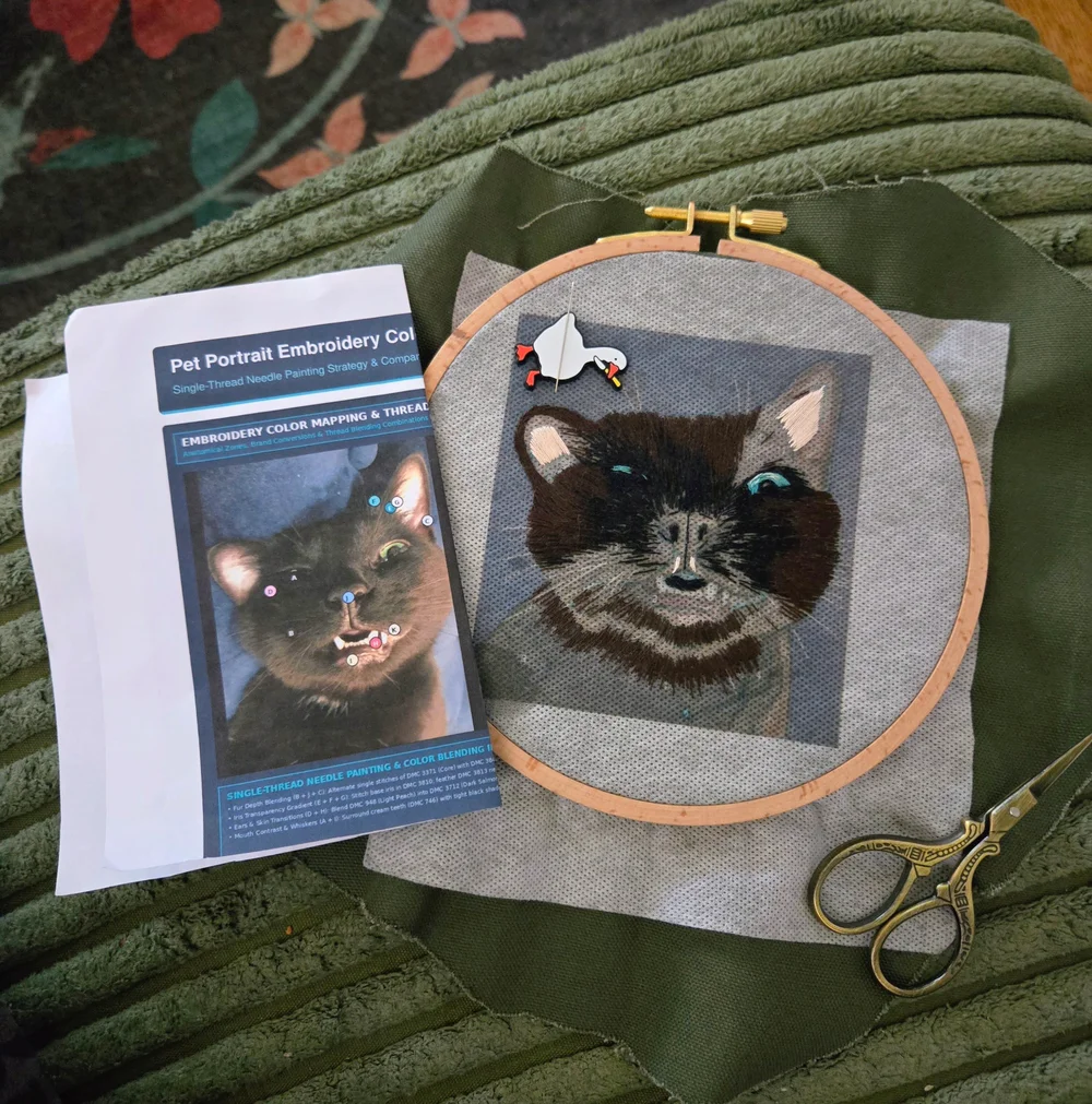 A black cat portrait being hand-embroidered in a wooden hoop beside its printed reference and embroidery scissors
